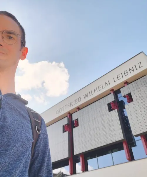 Eine männlich-gelesen Persönlichkeit steht draußen an einem sonnigen Tag vor einem Gebäude. Die Persönlichkeit trägt eine Brille und ist zur Hälfte zu sehen von Kopf bis zum Oberkörper. Dahinter das Gebäude mir der Schrift Gottfried Wilhelm Leibniz