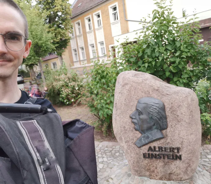 A personality, half-seen with glasses and a backpack, stands in front of and next to a public sculpture with his face in the profile of Albert Einstein. Below is the name.