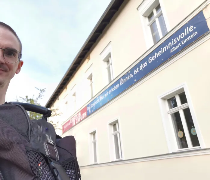 A figure, half-seen wearing glasses and a backpack, stands in front of a building with a banner. On the left, in white lettering against a red background, is the Einstein Exhibition in Caputh. To the right of it in the same font color with a blue background: The most beautiful thing we can experience is the mysterious. Albert Einstein